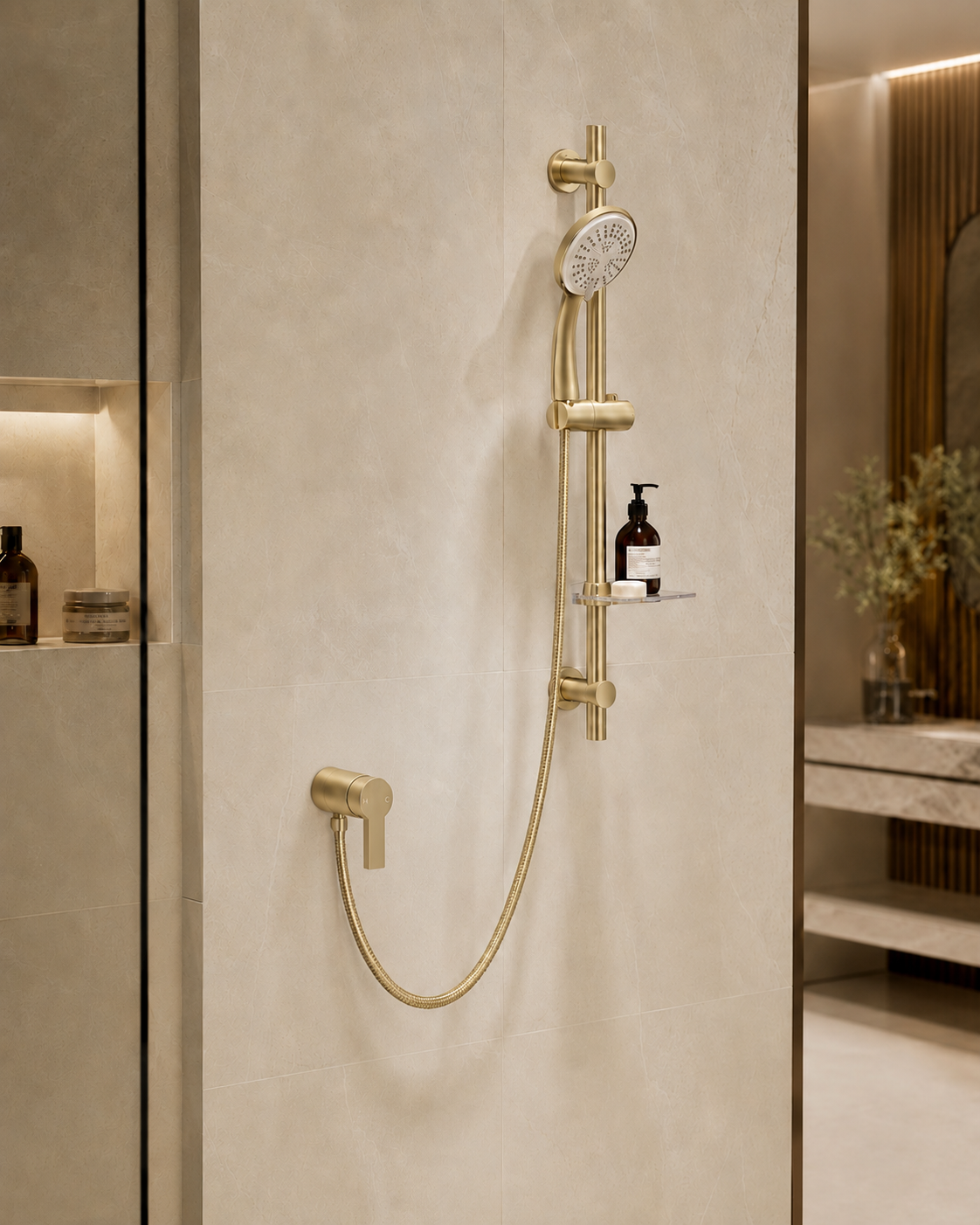 A modern wall-mounted shower fixture in a warm brushed brass or gold finish, installed on a beige tiled bathroom wall. The set includes a round shower head, flexible hose, wall-mounted control valve, and a small integrated shelf holding a soap dispenser.