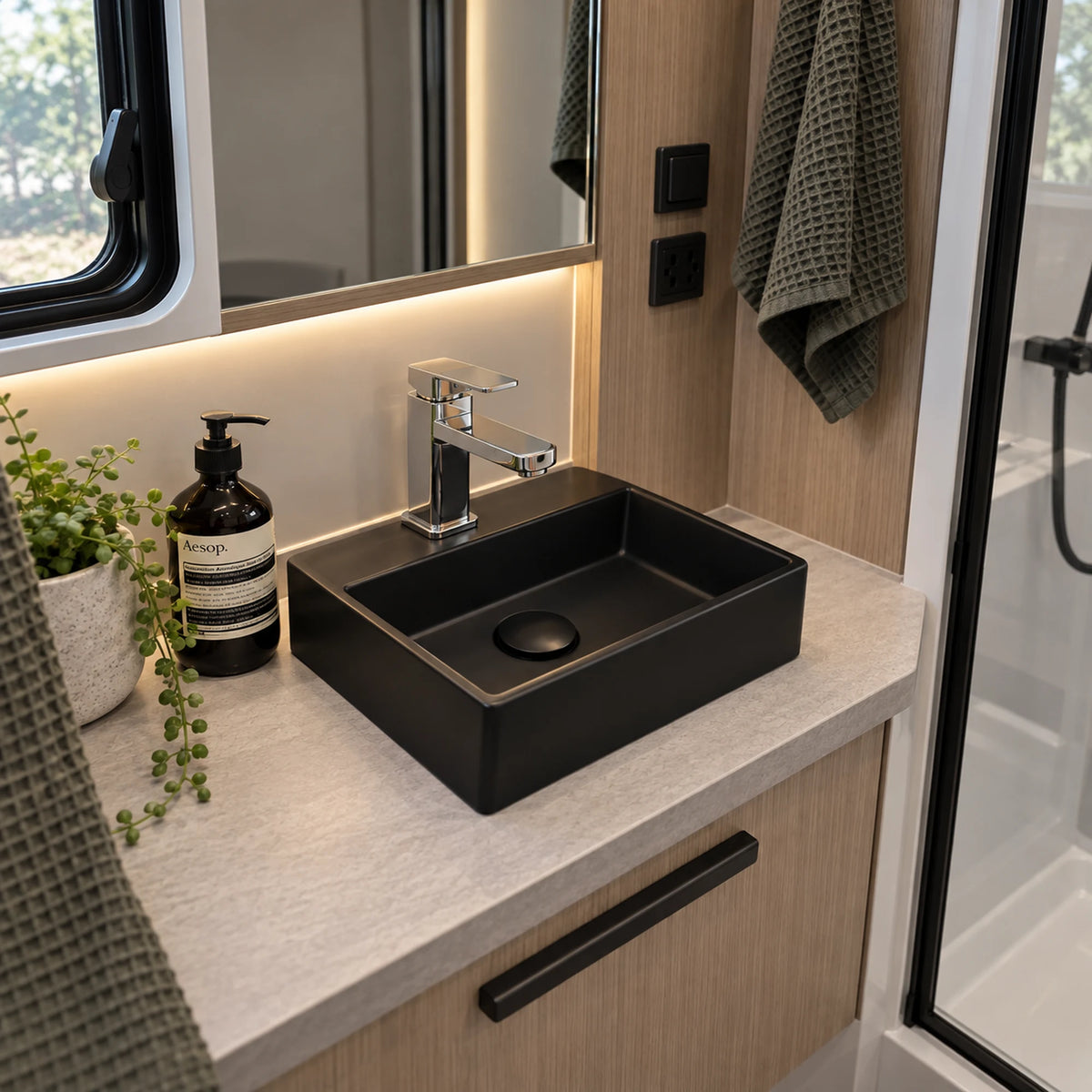 A modern matte black rectangular vessel sink with a chrome faucet, installed on a light stone countertop inside a compact bathroom or RV setup. A dark soap dispenser, small potted plant, wood-look cabinet, mirror, and shower glass are visible around it.